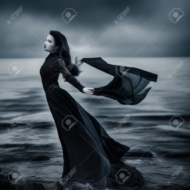 Hermosa chica gótica triste con un trapo en las manos de pie en la orilla del mar