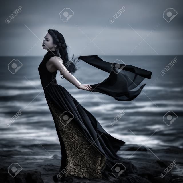 Beautiful sad goth girl with cloth in hands standing on the sea shore