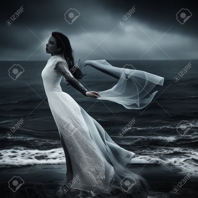 Hermosa chica gótica triste con un trapo en las manos de pie en la orilla del mar