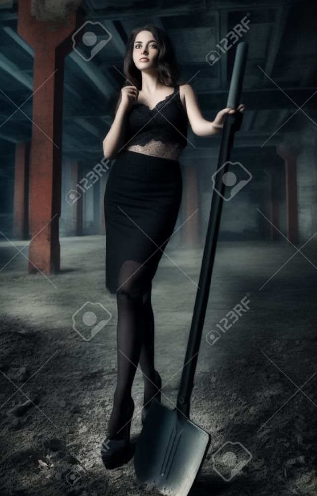 Portret van mooi meisje met schop in handen. Elegante jonge vrouw gekleed in zwarte rok en beha's onder verlaten industriële plaats