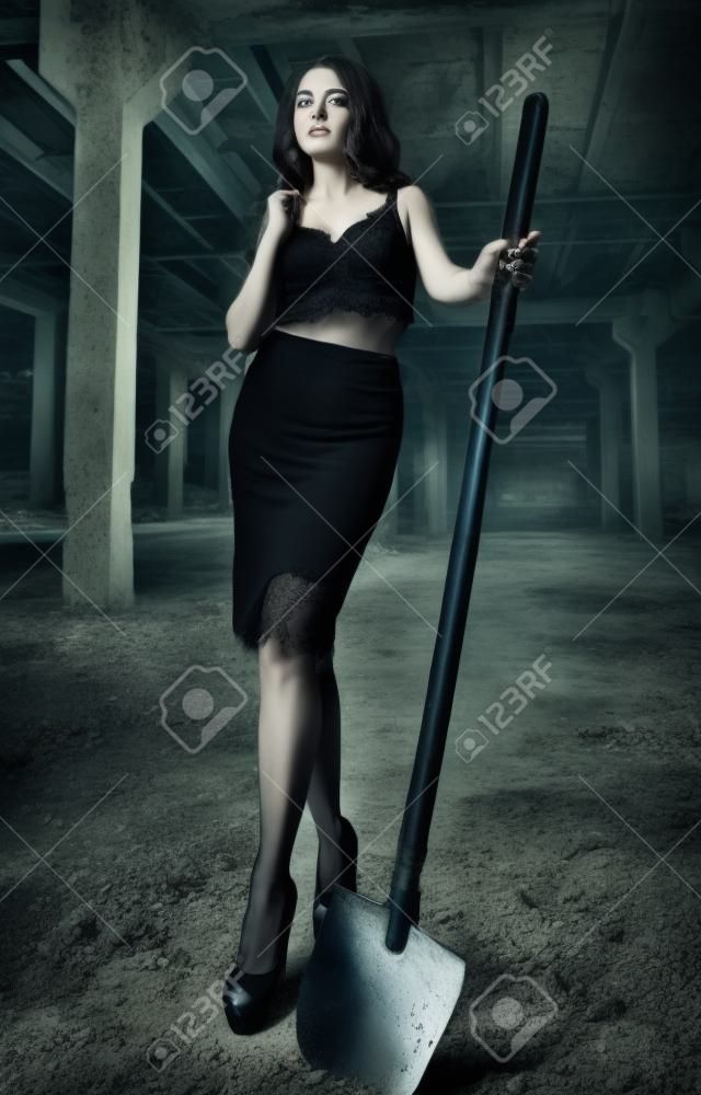 Portret van mooi meisje met schop in handen. Elegante jonge vrouw gekleed in zwarte rok en beha's onder verlaten industriële plaats