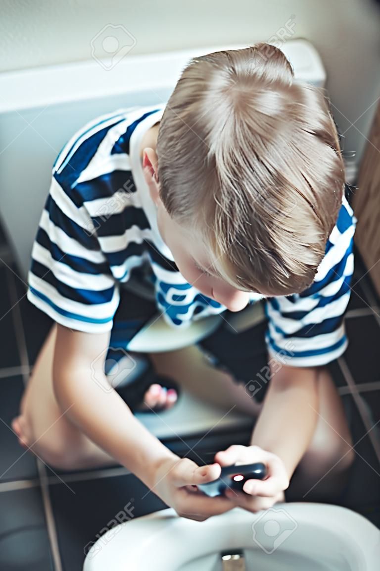 Nastoletniego chłopaka obsiadanie na toalecie z jego telefonem komórkowym w jego ręce sprawdza jego wiadomości tekstowe