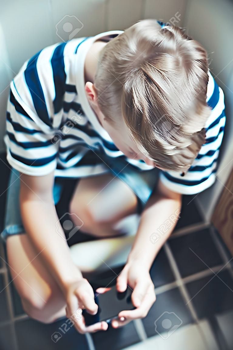 Nastoletniego chłopaka obsiadanie na toalecie z jego telefonem komórkowym w jego ręce sprawdza jego wiadomości tekstowe