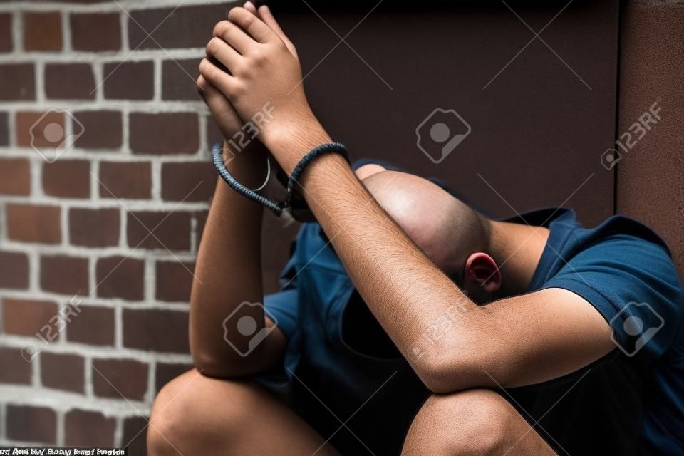 Dejected teenage boy held captive in handcuffs sitting on the ground against an exterior wooden door with his head down between his legs after being busted and arrested