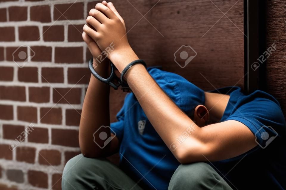 Dejected teenage boy held captive in handcuffs sitting on the ground against an exterior wooden door with his head down between his legs after being busted and arrested