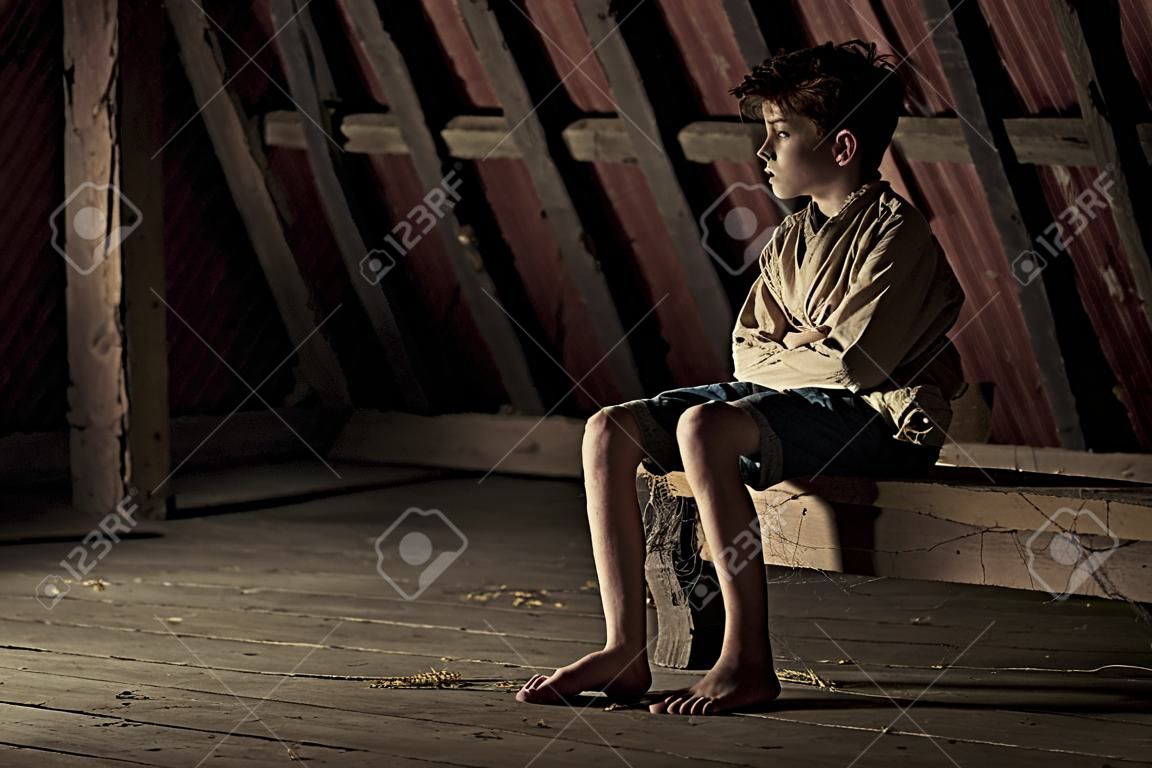 Barefoot tiener jongen geïmmobiliseerd in een rechte jas zittend op een houten balk bedekt met spinnenwebben op een schaduwrijke zolder in een conceptuele atmosferische afbeelding met kopieerruimte
