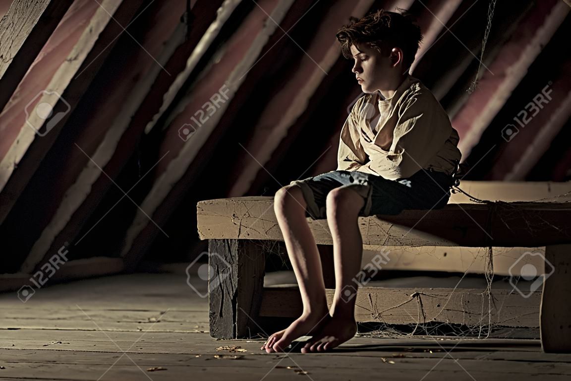 Barefoot tiener jongen geïmmobiliseerd in een rechte jas zittend op een houten balk bedekt met spinnenwebben op een schaduwrijke zolder in een conceptuele atmosferische afbeelding met kopieerruimte
