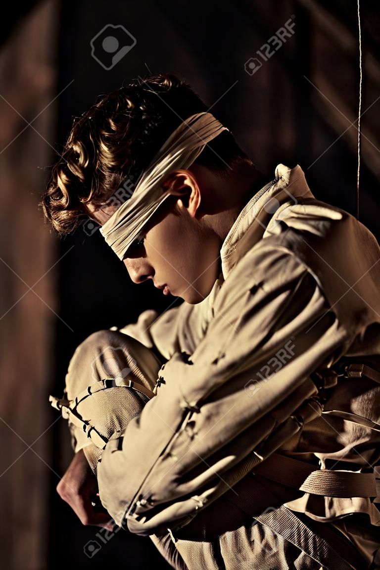 Junger Mann in einer zurückhaltenden oder geraden Jacke und Augenbinde in einem rustikalen Gebäude in Schatten in einem konzeptionellen Bild gebeugt