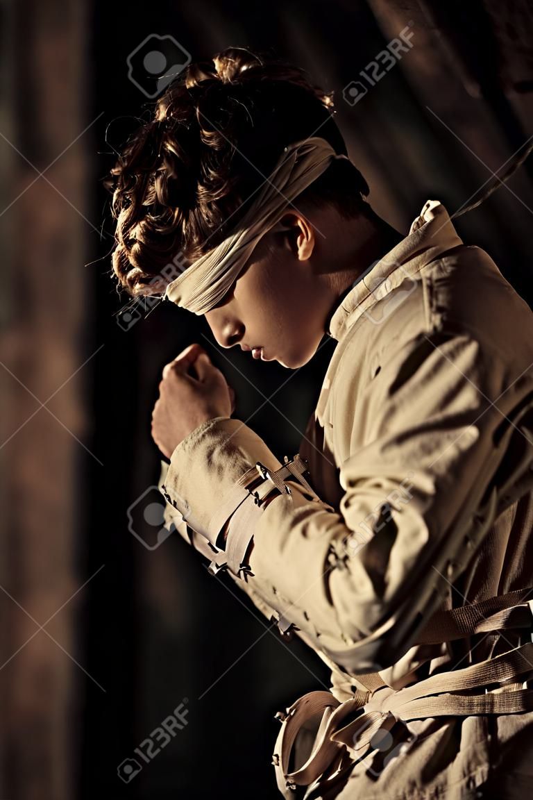 Junger Mann in einer zurückhaltenden oder geraden Jacke und Augenbinde in einem rustikalen Gebäude in Schatten in einem konzeptionellen Bild gebeugt