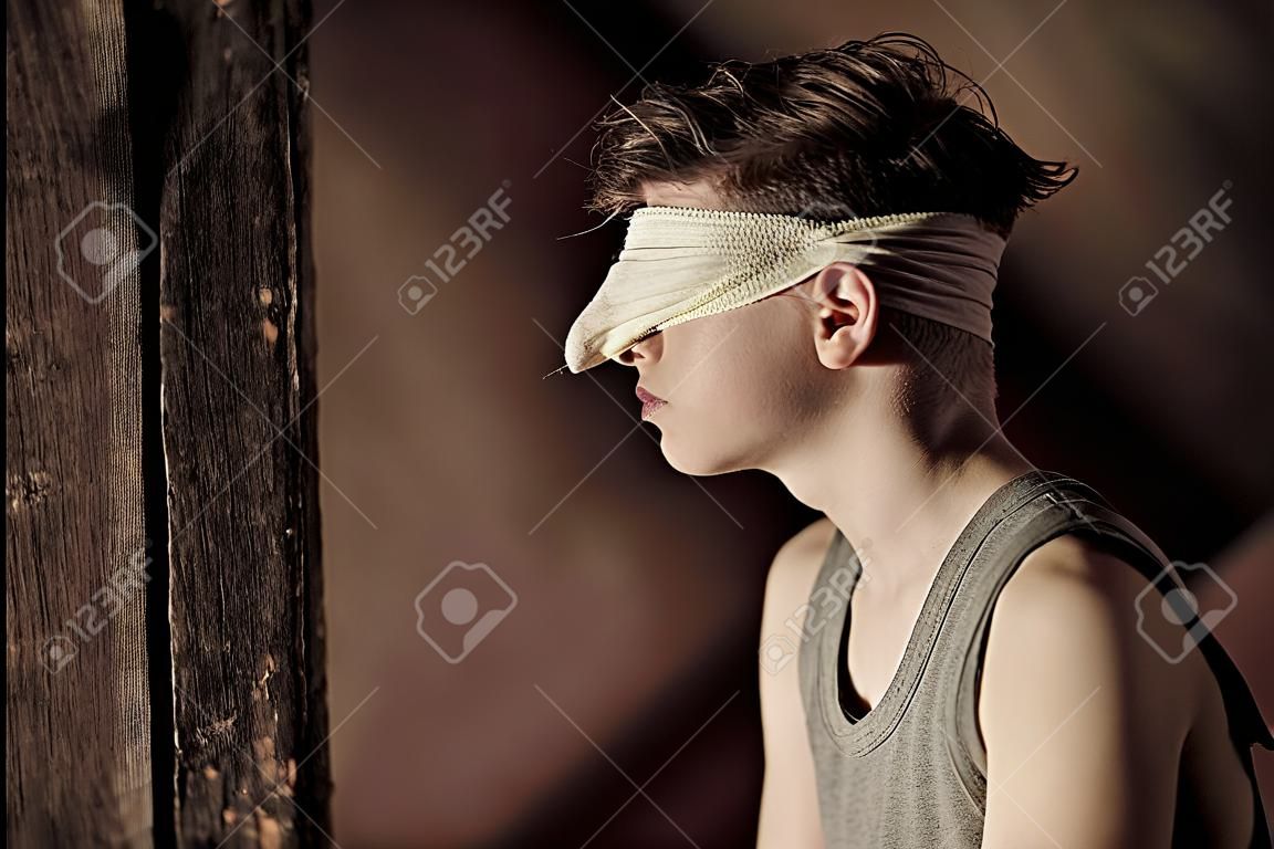 Teenage boy tied up in a blindfold sitting in an attic in the darkness in a conceptual image of abuse and hostage taking