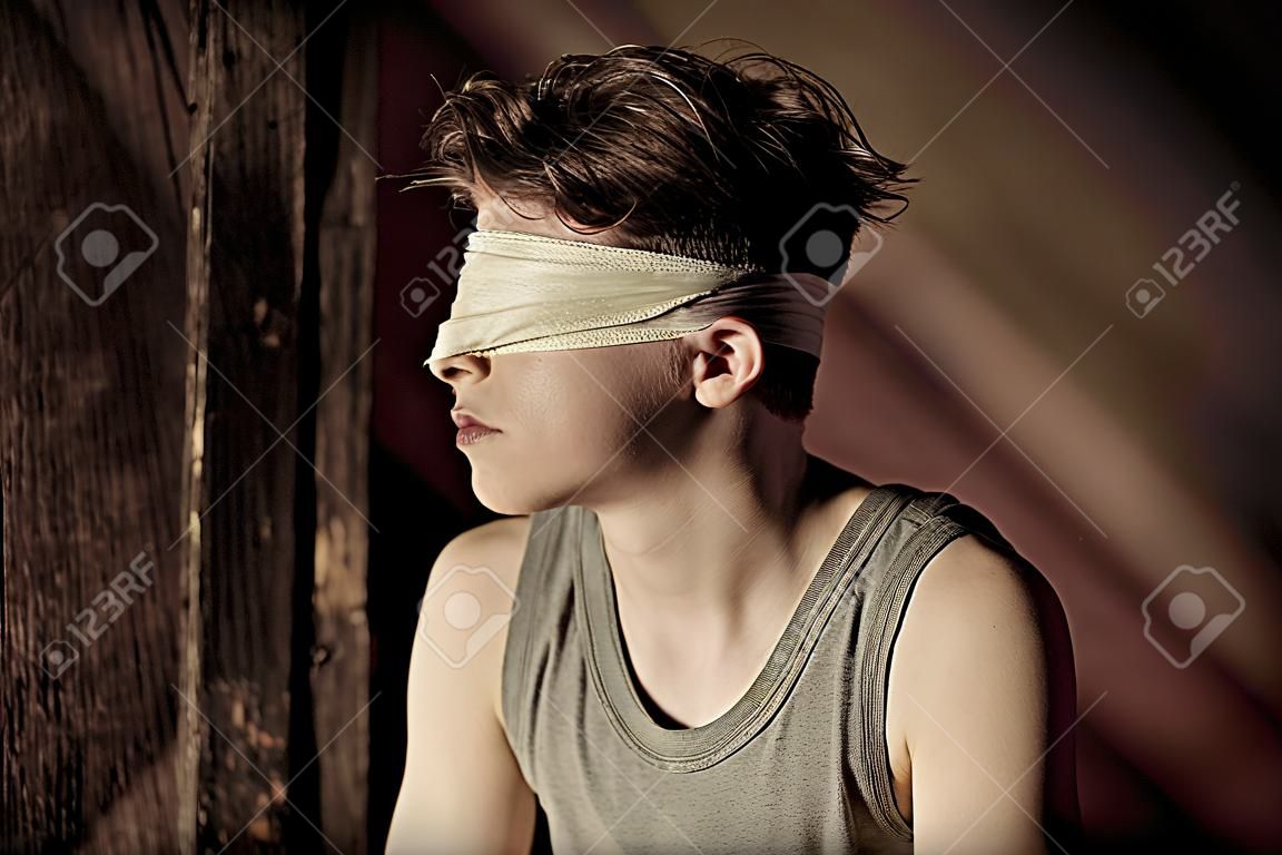 Teenage boy tied up in a blindfold sitting in an attic in the darkness in a conceptual image of abuse and hostage taking