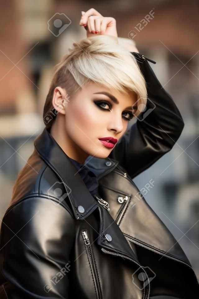 Young pretty stylish girl in black leather jacket posing, walking outdoors. Fashionable blonde woman with short hair looking at you