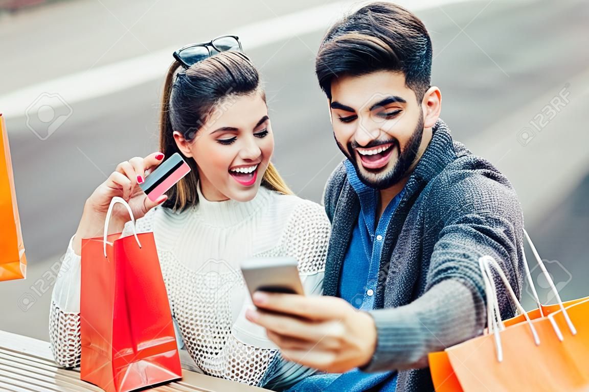 Bild von jungen Paaren mit Einkaufstaschen und Kreditkarte in der Stadt, die intelligentes Telefon, Online-Zahlung hält.