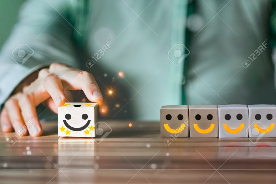Ręka biznesmena przesuwa ikonę uśmiechniętej twarzy z 5-gwiazdkowym zadowoleniem na drewnianym bloku sześcianowym, przedstawiając koncepcję obsługi klienta, najlepszą, doskonałą ocenę biznesową
