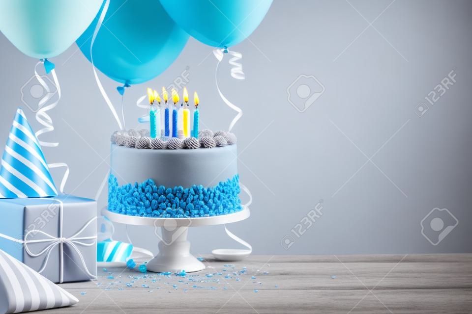 Açık renkli gri üzerinde mavi doğum günü pastası, hediyeler, şapkalar ve renkli balonlar.