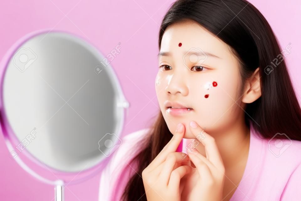 Asiatische Teenager-Frau, die Spiegel betrachtet und Akne-Problem auf ihrem Gesicht zusammendrückt, Hautpflegekonzept.