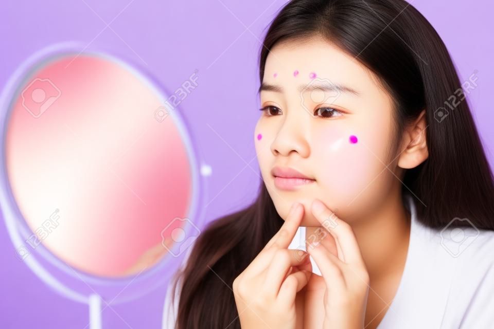 Asiatische Teenager-Frau, die Spiegel betrachtet und Akne-Problem auf ihrem Gesicht zusammendrückt, Hautpflegekonzept.