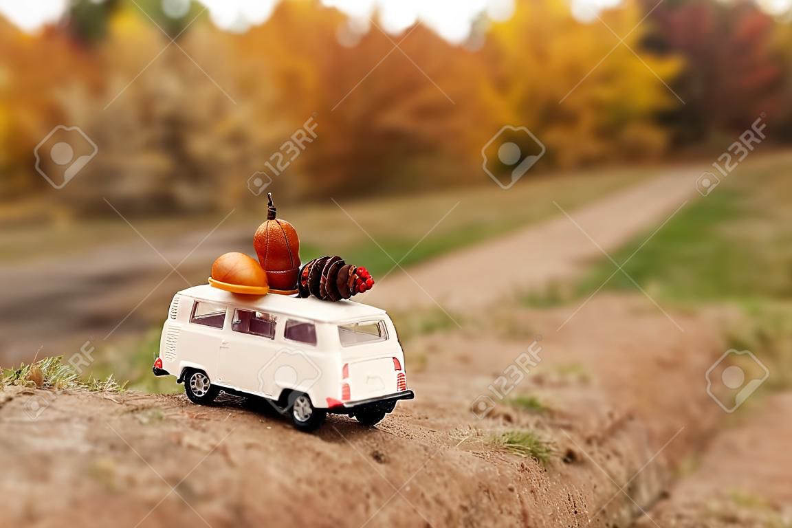 miniatuur speelgoed auto minivan draagt op het dak gele bladeren, eikel, kegel en rode bessen op de achtergrond van de herfst natuur en bomen.
