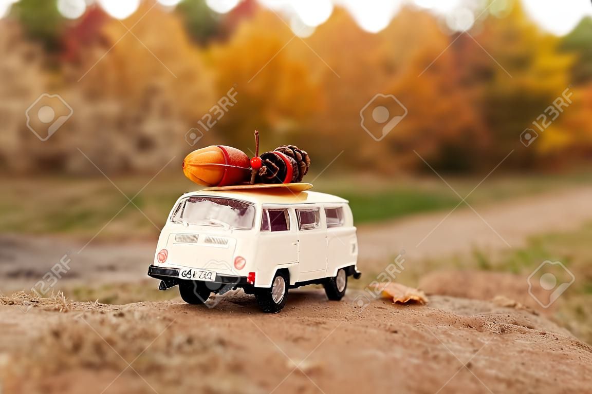 miniature toy car minivan carries on the roof yellow leaves, acorn, cone and red berries on the background of autumn nature and trees.