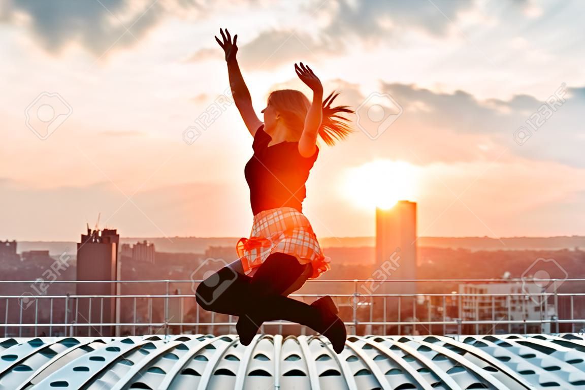 Siluetta della donna allegra felice che salta e che si diverte in città contro il tramonto. Concetto di vacanza di libertà e svago.