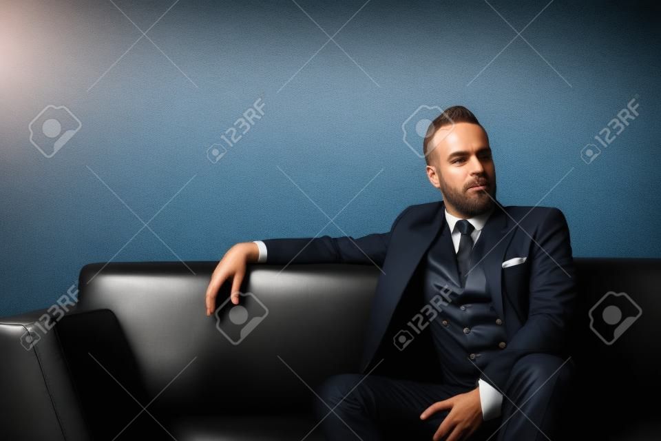 Portrait of adult businessman wearing trendy suit and sitting modern studio on leather sofa against the empty wall.