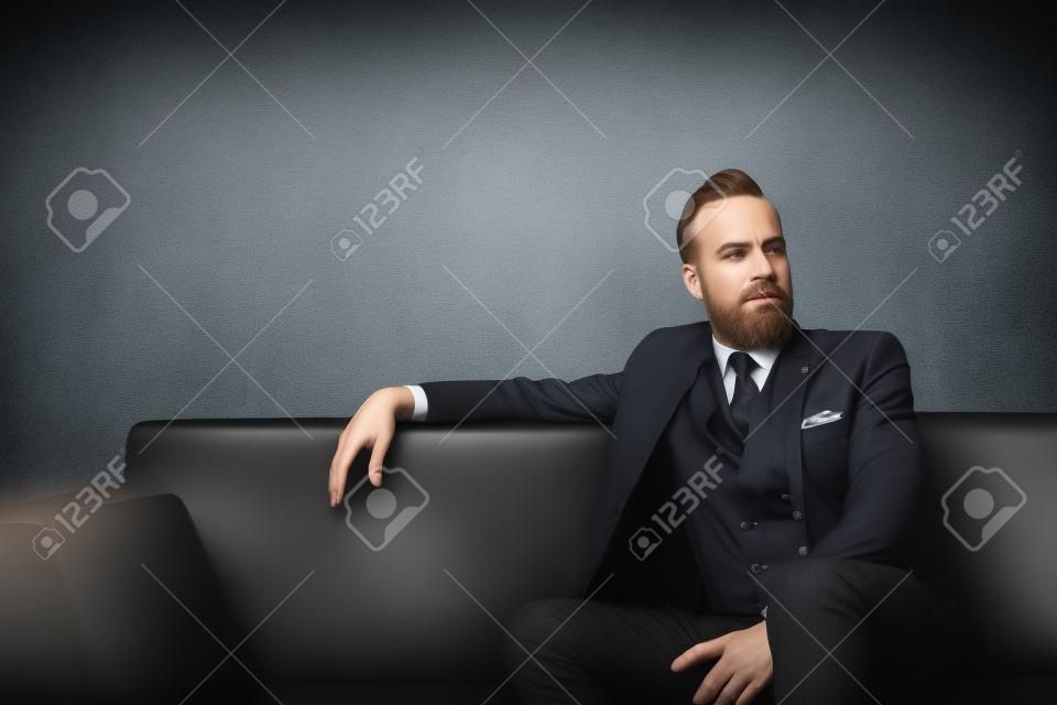 Портрет взрослых бизнесмен носить модный костюм и сидит современная студия на кожаный диван на фоне пустой стены.