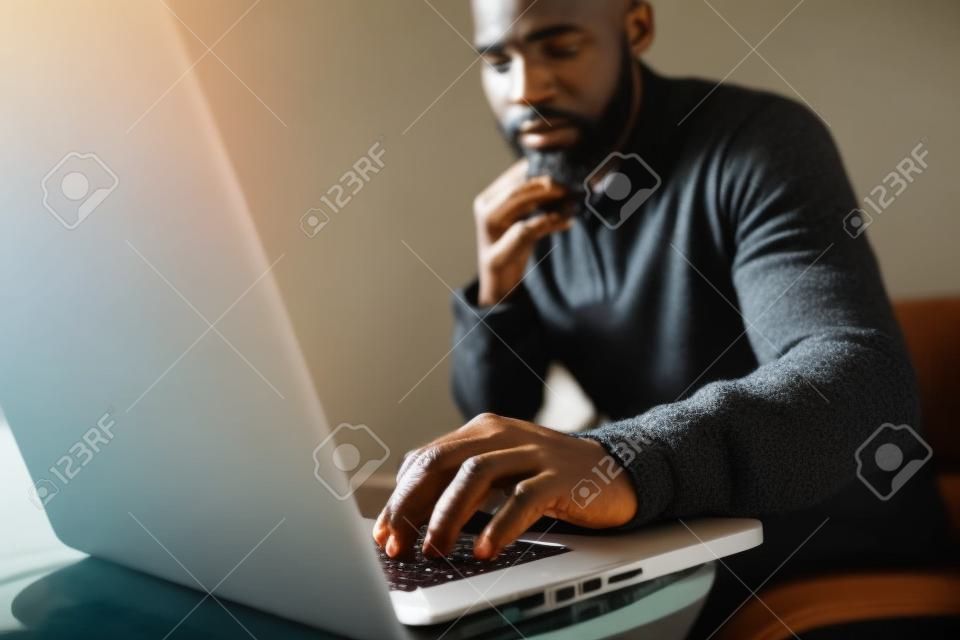 Attrayant homme africain barbu utilisant un ordinateur portable tout en étant assis sur le canapé à son bureau à la maison moderne. Concept de jeunes bénéficiant d'appareils mobiles. Gros plan avec une main masculine de mise au point.