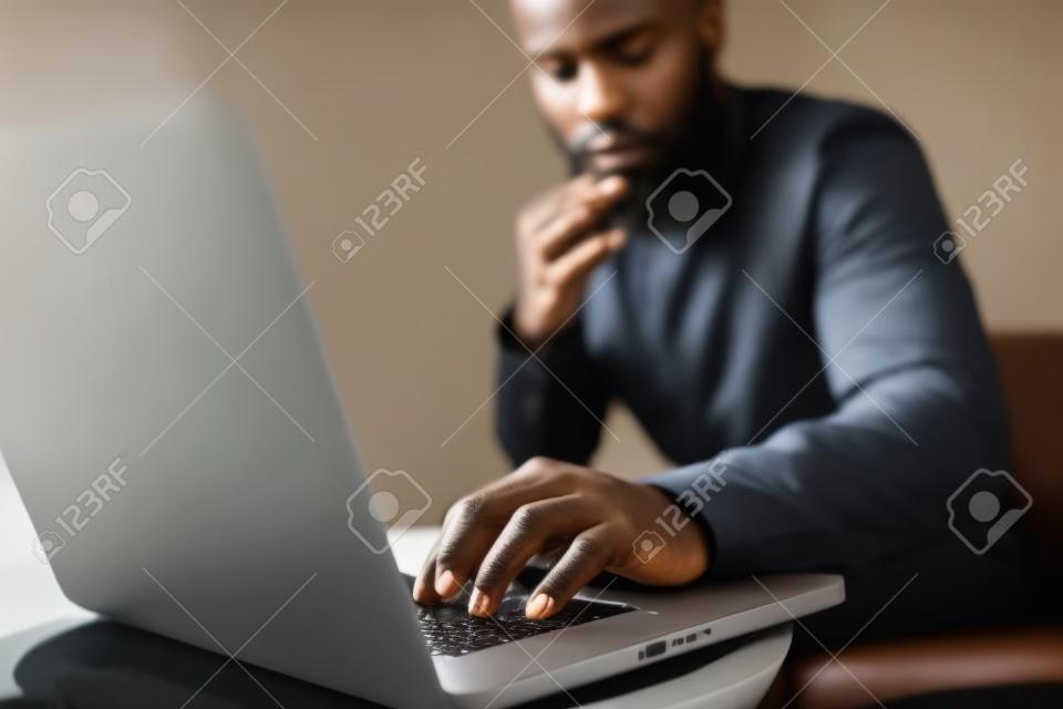 Attrayant homme africain barbu utilisant un ordinateur portable tout en étant assis sur le canapé à son bureau à la maison moderne. Concept de jeunes bénéficiant d'appareils mobiles. Gros plan avec une main masculine de mise au point.