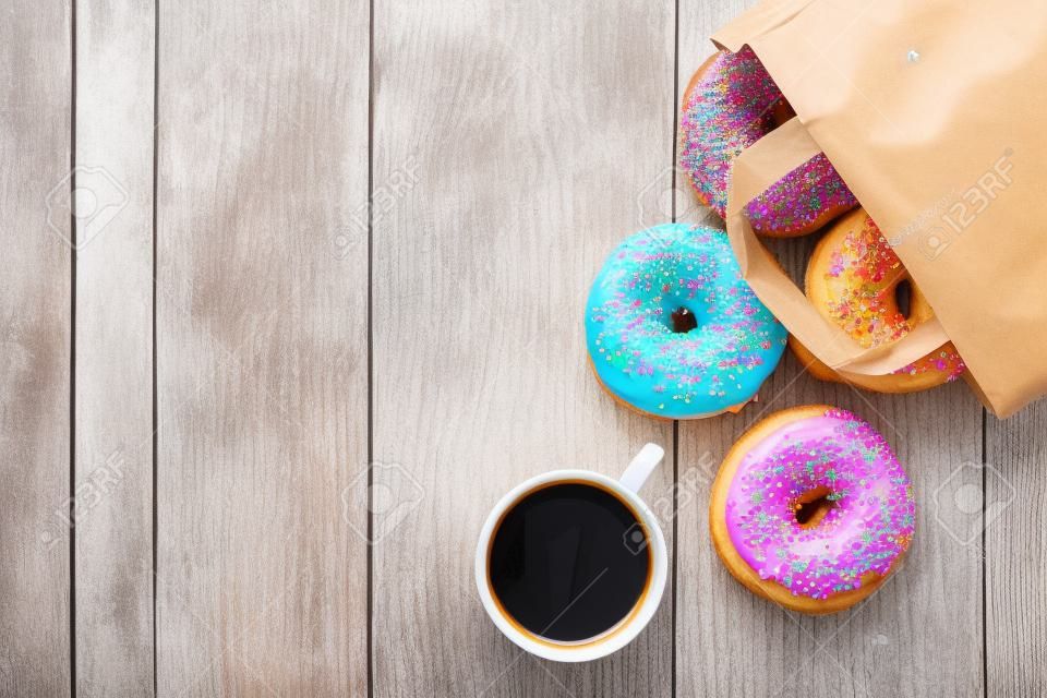 Bunte Donuts in Papiertüte und Kaffeetasse auf Holztisch. Ansicht von oben mit Kopie Raum
