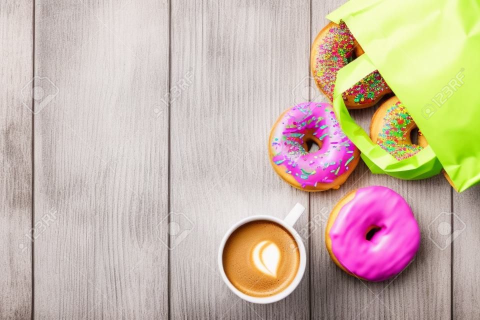 Bunte Donuts in Papiertüte und Kaffeetasse auf Holztisch. Ansicht von oben mit Kopie Raum