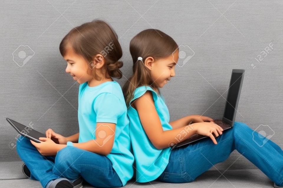 Dos niños sentados espalda con espalda socializar con ordenador portátil y tablet.