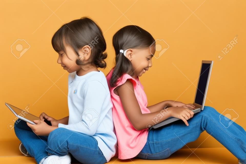 Dos niños sentados espalda con espalda socializar con ordenador portátil y tablet.