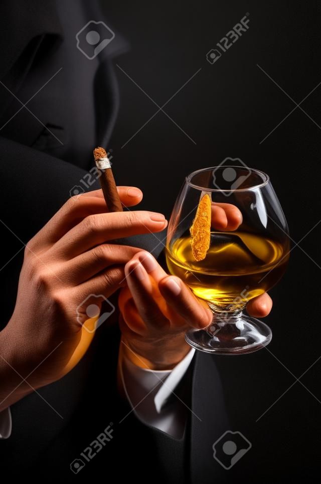 在男性手上的旧白兰地酒杯，在黑色背景下抽雪茄