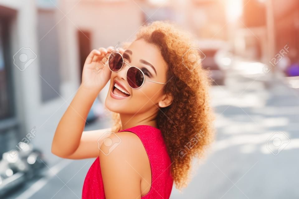 Mujer joven en gafas de sol redondas y vestido con el pelo rizado sonriente sobre el hombro en la calle de la ciudad