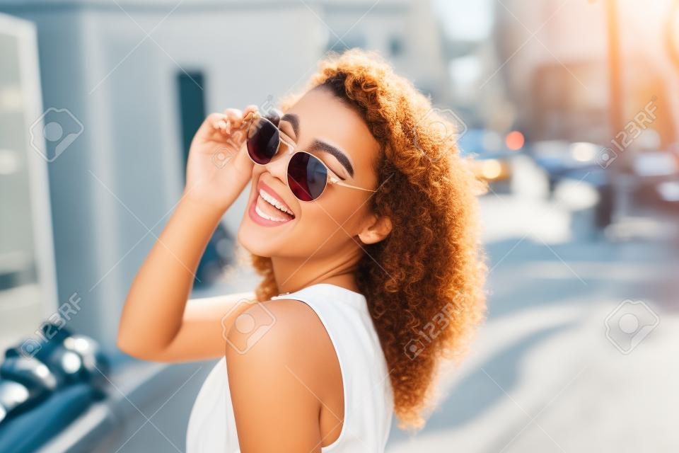 Mujer joven en gafas de sol redondas y vestido con el pelo rizado sonriente sobre el hombro en la calle de la ciudad