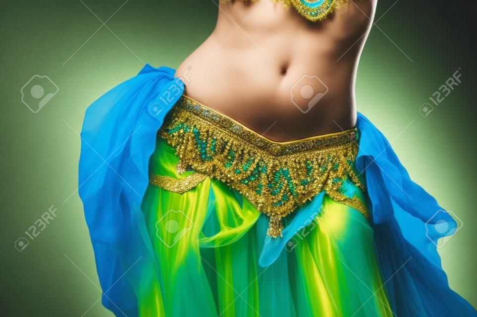 Close up shot of a belly dancer wearing a blue, gold and green costume shaking her hips  

