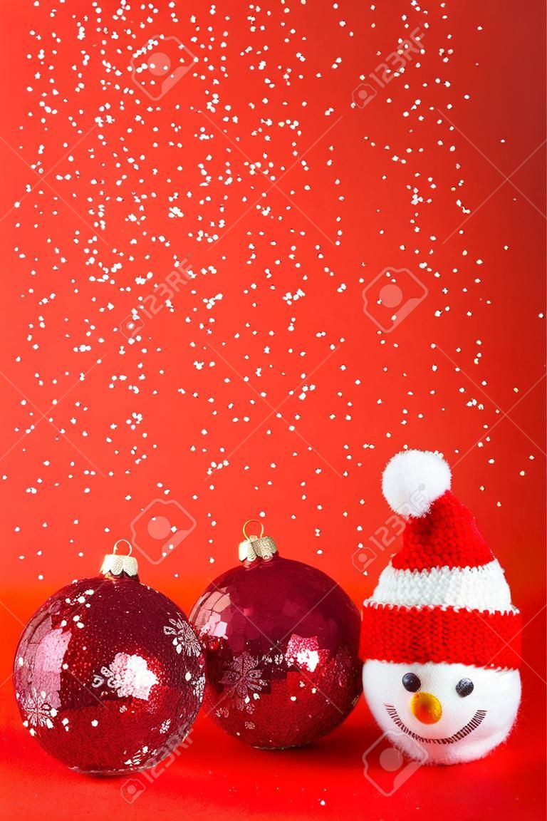 Knitted snowman in a red Christmas hat and sweater with two red balls on a red background with falling snow. Merry Christmas and New Year 2023 greeting card