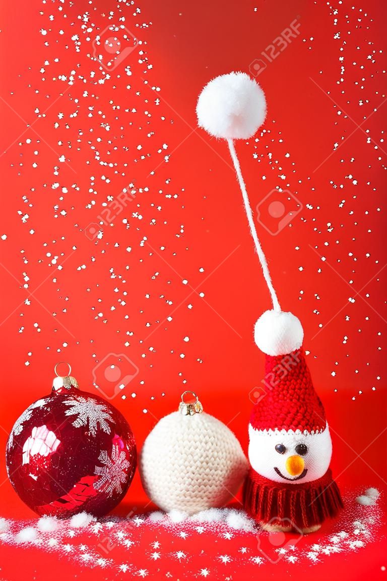 Gestrickter Schneemann mit roter Weihnachtsmütze und Pullover mit zwei roten Kugeln auf rotem Hintergrund mit fallender Schnee-Grußkarte für frohe Weihnachten und das neue Jahr 2023