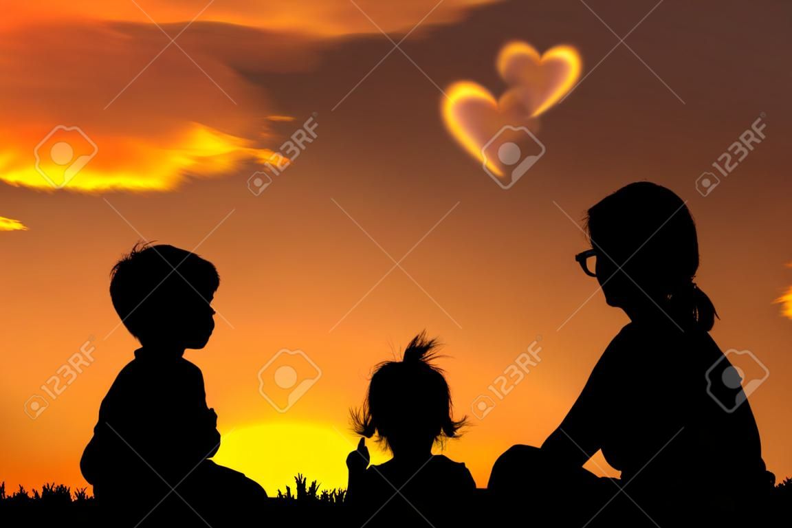 Силуэт матери и двух детей сидит и смотрит небо на закате. Маленькая точка мальчик сердца формы облаков. Красочный фон неба оранжевый и золотой цвета закатного неба. Дружная семья.
