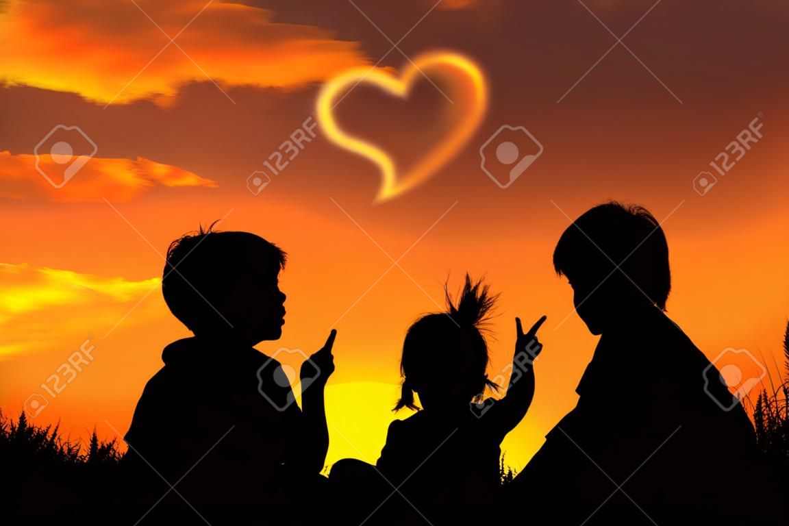 Silueta de la madre y dos niños sentados y mirando el cielo al atardecer. Niño pequeño punto a las nubes en forma de corazón. Fondo colorido del cielo cielo anaranjado y de los colores oro de la puesta del sol. Familia amable