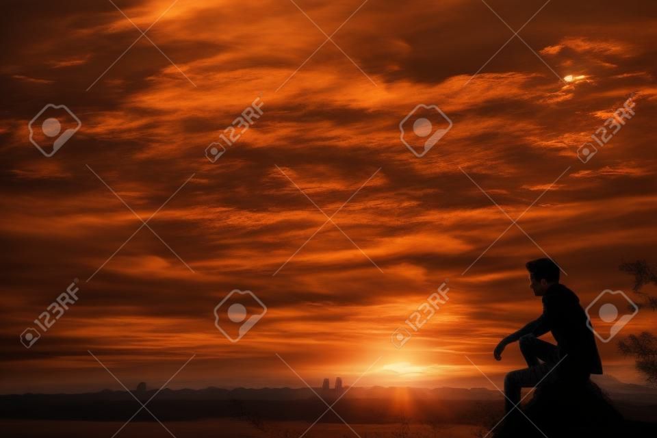 Силуэт молодого человека, сидящего на закате или восходе солнца. Уверенный подросток thinkig на скале камня. Надеюсь. Печаль. Свобода.