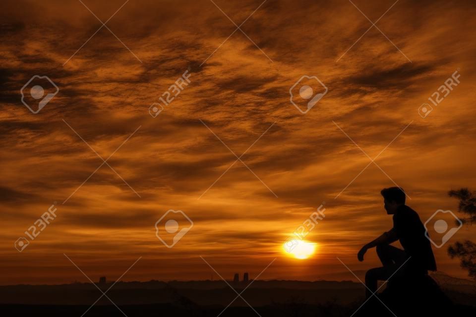 Silhouette of young man sitting on sunset or sunrise. Confident teenage boy thinkig on cliff stone. Hope. Sadness. Freedom.