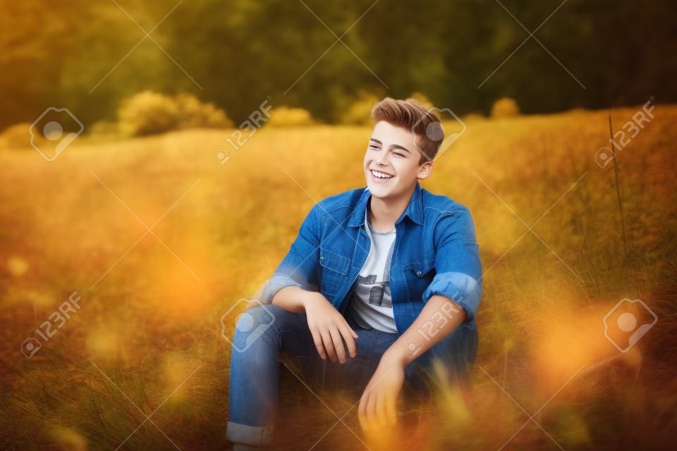 Knappe tiener jongen draagt casual shirt zittend op gras op de herfst dag. Gelukkige glimlachende jongeman zitten buiten.