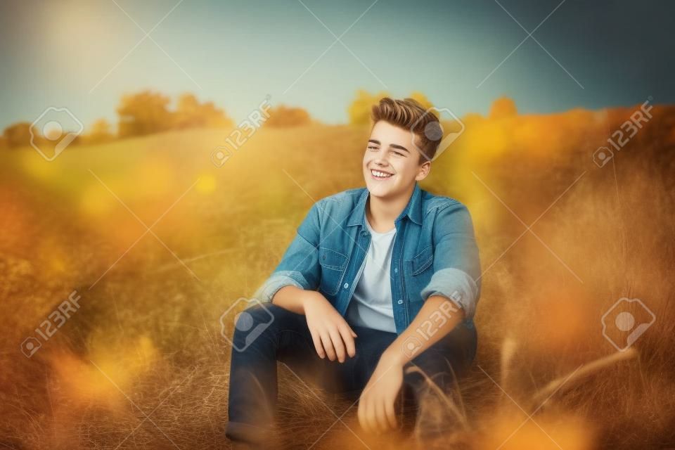 英俊的少年穿著休閒襯衫坐在草地上的秋日。幸福的微笑的年輕男子坐在戶外。