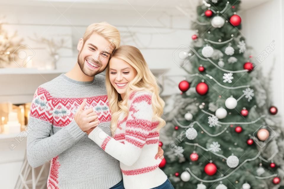 Casal doce em suéteres de inverno abraçando-se no fundo de decorações de Natal. Homem bonito e mulher jovem loira celebrando as férias de inverno