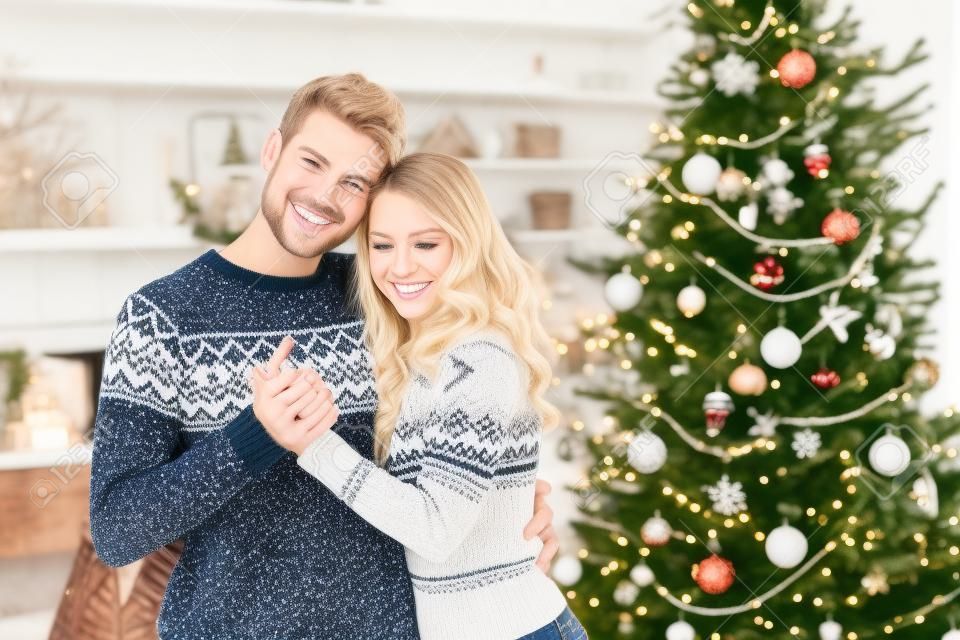 Casal doce em suéteres de inverno abraçando-se no fundo de decorações de Natal. Homem bonito e mulher jovem loira celebrando as férias de inverno