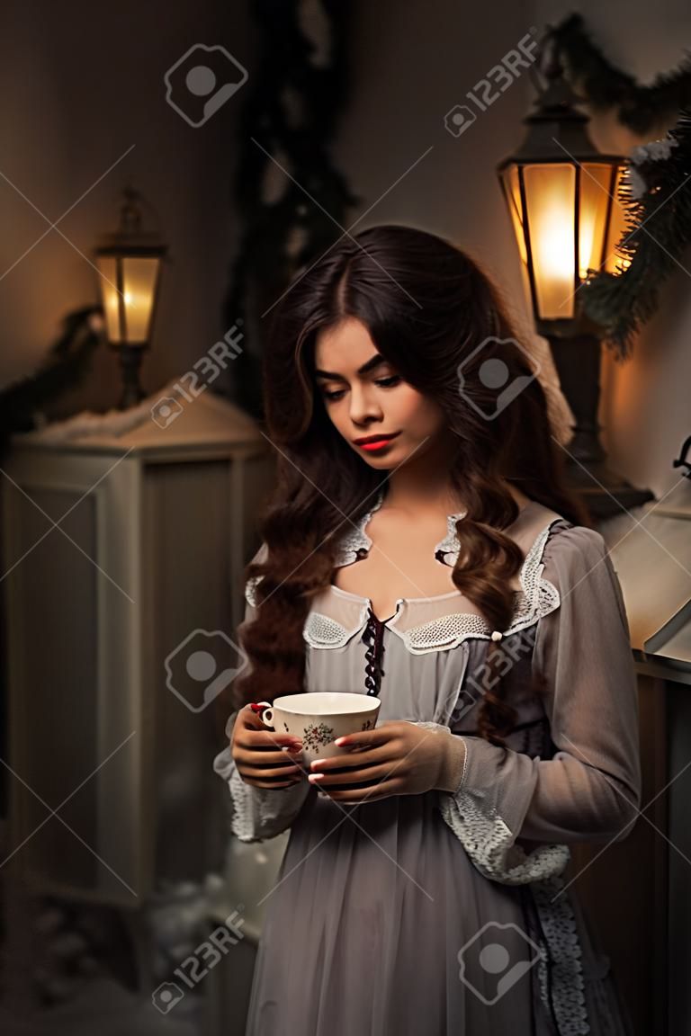 The Winter's Tale. Mooie vrouw in vintage jurk staan naast het huis met een kopje thee. Lang haar, baby gezicht. Creatieve kleuren