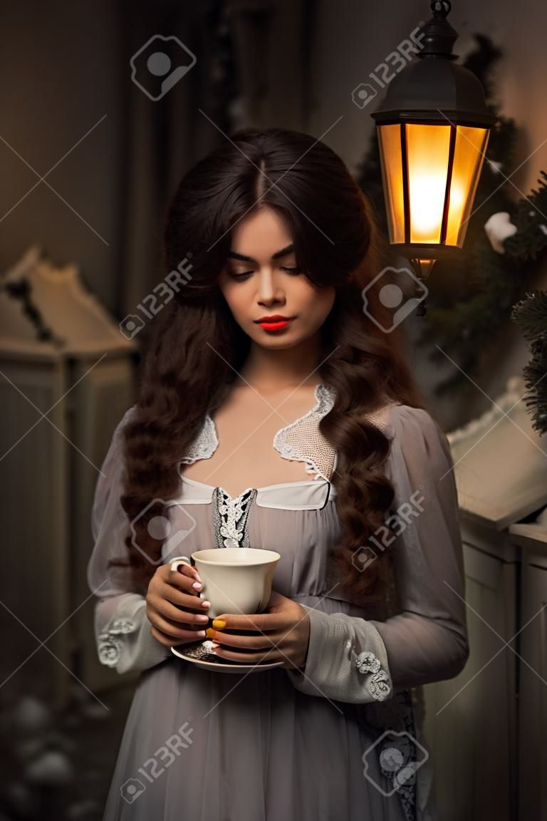 Зимняя сказка. Красивая женщина в винтажном платье, стоя рядом с домом с чашкой чая. Длинные волосы, детское лицо. Творческие цвета