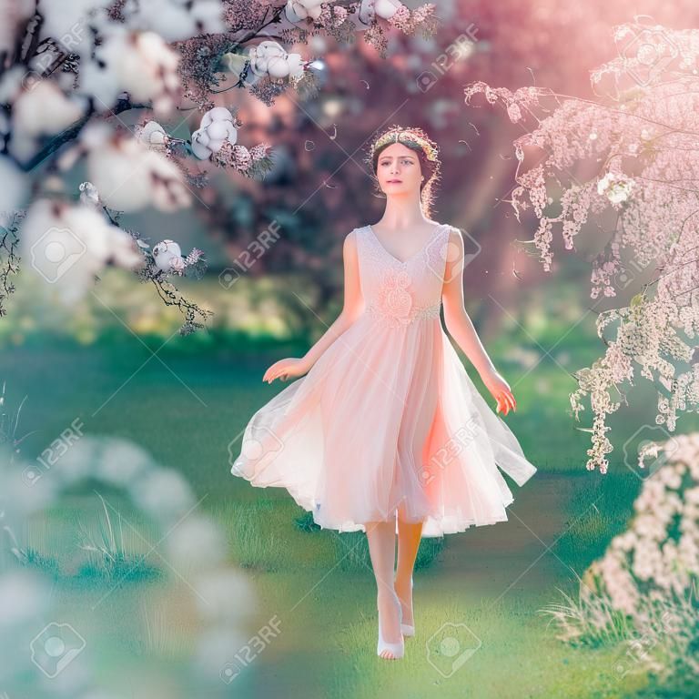mysterieuze dame met donker verzameld haar in een licht delicate perzik jurk loopt in de bloeiende tuin, de lente fee raakt haar bezittingen, de sprookjes elf als een pop op het gras blootsvoets