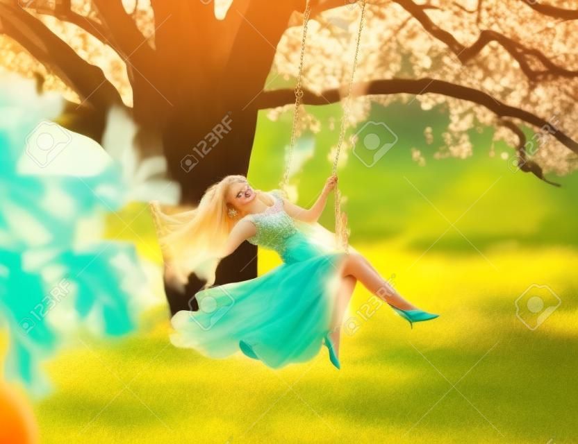 Mooie gelukkige vrouw nimf zitten op schommels. Magische fantasie schommel. prinses lange perzik kleur oranje zijde vintage jurk uitwaaierende wind. blond haar vliegen in beweging. Boom zonneschijn groen gras bos
